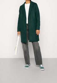 Dunkelgrüner langer Mantel mit gekerbtem Revers und zwei Knöpfen, kombiniert mit grauen Jeans und grünen Turnschuhen. In der Hand wird eine weiße Umhängetasche gehalten.