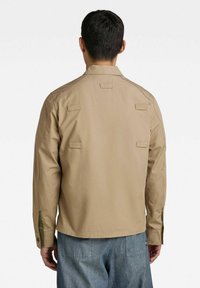 Giacca leggera color khaki con vestibilità rilassata, maniche lunghe e polsini con bottone. Presenta diversi dettagli con toppe sulla schiena.