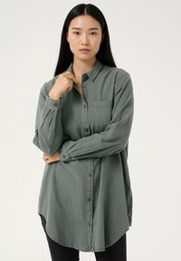 Hele roheline, pika varrukaga särk, mis on valmistatud tekstureeritud kangast. Esiosas nööbitav, rinnataskuga ja kaardus servaga. Kanditud tumevärviliste pükstega.