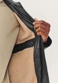 Main tenant une veste en cuir noire ouverte montrant la doublure en polaire beige et la poche intérieure sur un fond uni.