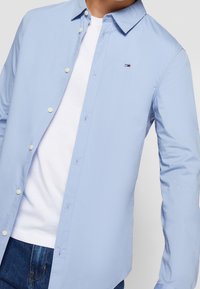 Uomo che indossa una camicia azzurra a maniche lunghe sbottonata sopra una maglietta bianca e jeans blu, con un piccolo logo ricamato sul petto.