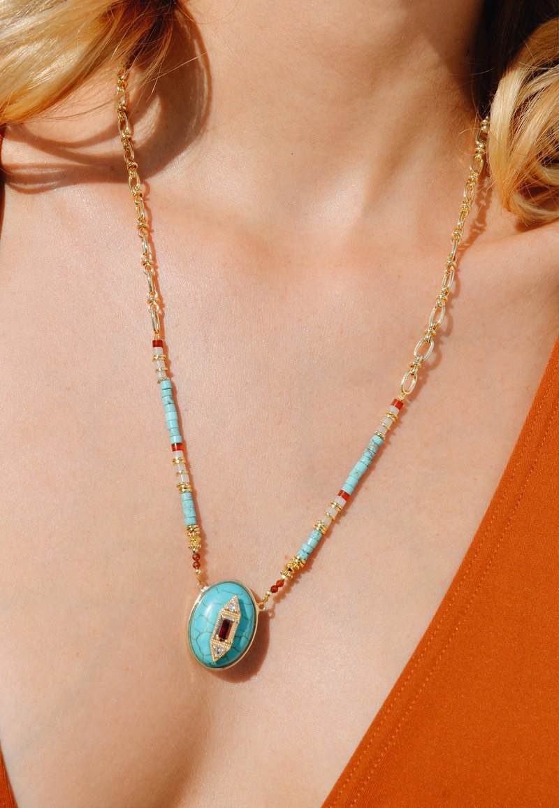 Collier en perles turquoise avec des accents dorés, présentant un pendentif ovale avec un détail en pierre rouge, sur fond de teint.