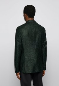 Mörkgrön kavaj, strukturerad tyg, skräddarsydd passform, enkel slits, snedskurna slag, knäppta manchetter, minimalistisk design med subtil prickmönstring.