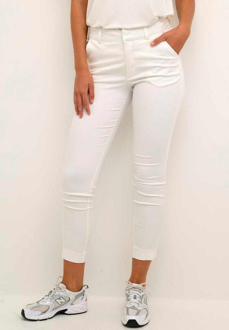 Pantalons blancs ajustés en tissu extensible, avec une taille mi-haute et des poches latérales, associés à des chaussures de sport gris et blanc.