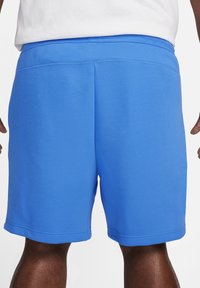 Bakifrån en person som bär ljusblå knä-långa shorts och en vit skjorta, står med armarna avslappnade vid sidorna.