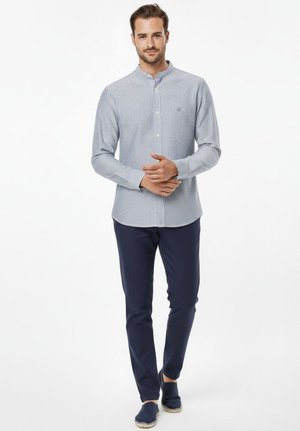 Hombre vestido con camisa de cuello mao a rayas azul claro, pantalones azul marino y zapatillas azul marino, posando frente a un fondo blanco.