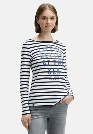 Junge Frau mit kurzen Haaren, die ein langärmliges schwarz-weiß gestreiftes Shirt mit blauer Schrift "Das Leben ist besser am Meer" und grauen Jeans trägt.