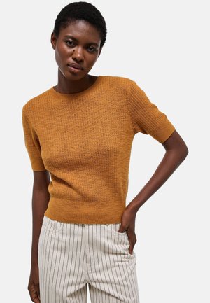 Femme portant un pull moutarde texturé à manches courtes et un pantalon blanc à fines rayures verticales noires, posant avec une main dans la poche.