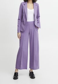 Lila kavaj med slagkrage, matchad med vida lila byxor. Båda är gjorda av ett texturerat tyg. Svarta loafers fulländar looken.