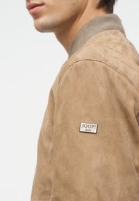 Νεαρός άνδρας που φοράει ένα ανοιχτό καφέ σουέντ μπουφάν με ραμμένη λεπτομέρεια στους ώμους και μεταλλικό λογότυπο "JOOP! JEANS" στο πάνω μέρος του μπράτσου.