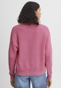 Rosa gestrickter Pullover mit lockerem Schnitt, strukturiertem Material, Rundhalsausschnitt und gerippten Bündchen sowie Saum. Kombiniert mit hellblauen Jeans.