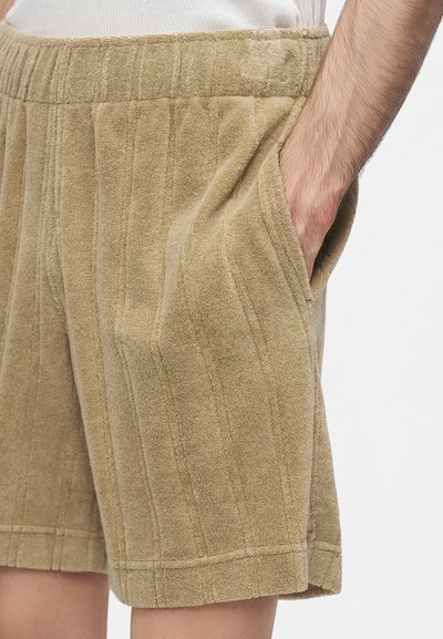 Gros plan d'une personne portant un short beige en tissu éponge avec une ceinture élastique, main reposant dans la poche droite sur fond blanc.