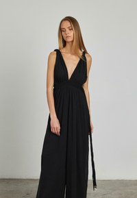 Vestido negro sin mangas que presenta un escote en V profundo, una cinturilla trenzada y una silueta fluida, con un detalle de lazo trenzado. Textura de tejido suave.