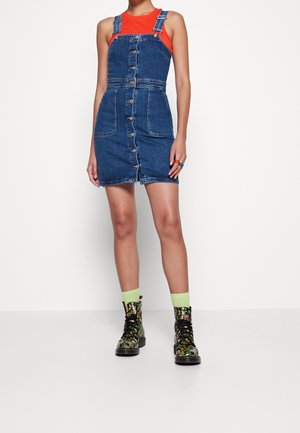Mujer con vestido peto de mezclilla con botones en la parte delantera sobre una camiseta sin mangas roja, botas con cordones florales y calcetines verde claro.