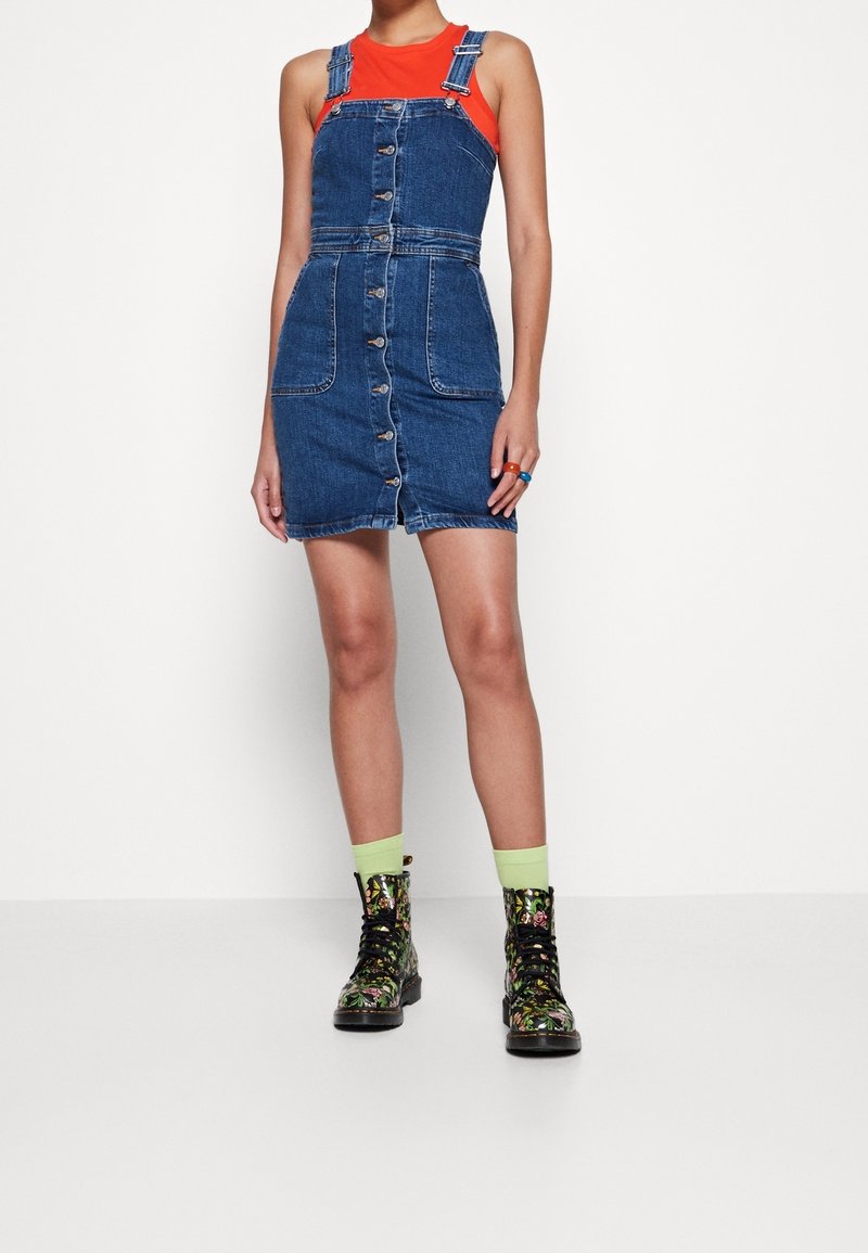 Femme portant une robe salopette en jean boutonnée sur le devant, un top rouge sans manches, des bottines lacées fleuries et des chaussettes vert clair.