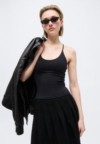 Νεαρή γυναίκα με μαύρο τοπ, μαύρη πλισέ φούστα, γυαλιά ηλίου και κρατώντας ένα μαύρο δερμάτινο μπουφάν στον ώμο της, μπροστά σε απλό φόντο.