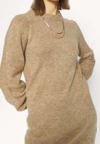 Beige neulepusero, jossa on pyöreä pääntie, pitkät hihat ja pehmeä rakenne. Asusteena kerroksellinen kultaketju.