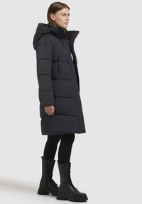 Puffjakke i mørk marineblå, med hætte, lynlås foran og store quiltede sektioner. Båret med sorte snørebåndsknæstøvler.