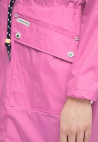 Vaaleanpunainen vedenpitävä takki, jossa on etutasku, nyörikiinnitys ja metalliset napit. Ominaisuuksiin kuuluu sileä, kiiltävä pinta.