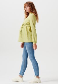 Blusa maternità verde chiaro, con texture e un nodo in vita, abbinata a jeans aderenti blu e sneakers bianche. Maniche lunghe e design casual.
