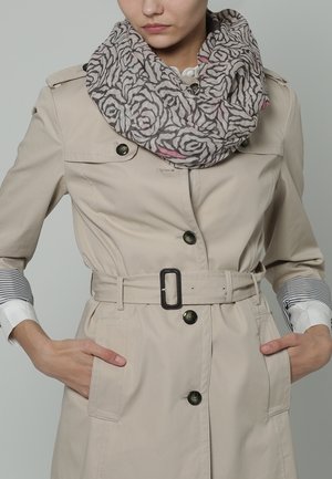 Femme portant un trench-coat beige ceinturé et une écharpe grise à motifs, les mains dans les poches, fond neutre.