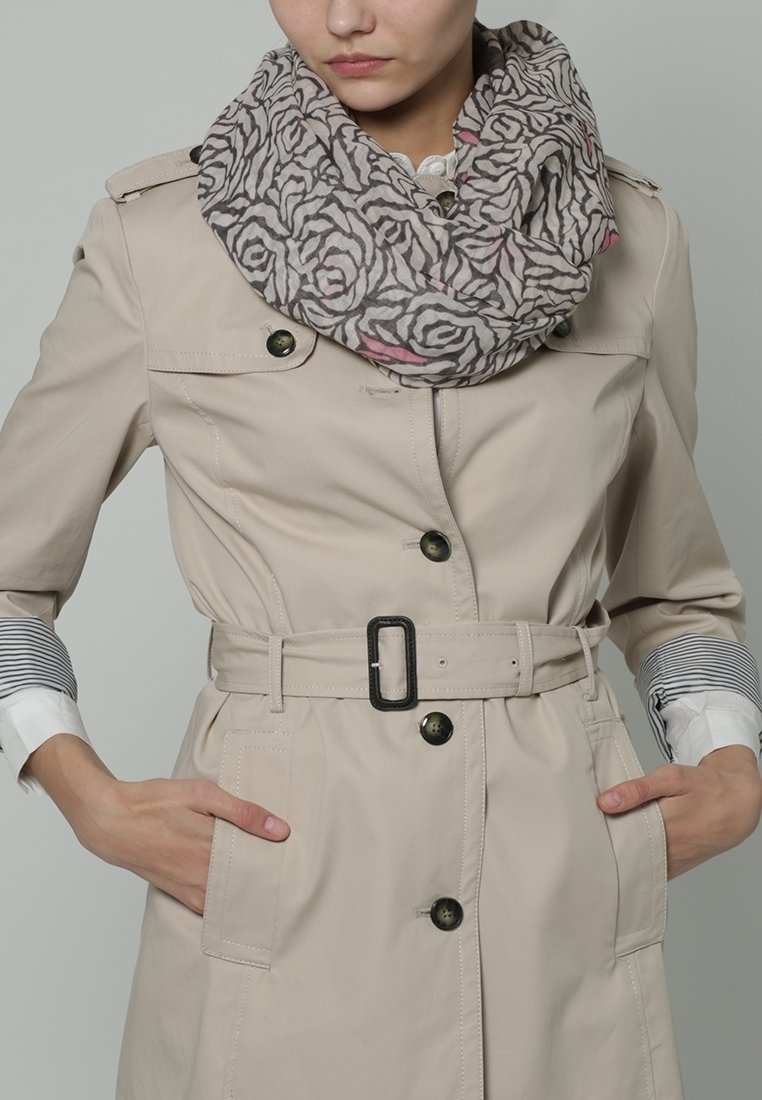 Femme portant un trench-coat beige ceinturé et une écharpe grise à motifs, les mains dans les poches, fond neutre.