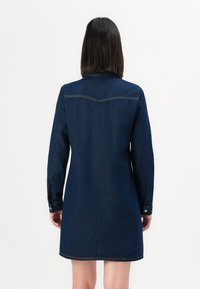 Femme portant une robe en denim bleu foncé à manches longues, se tenant de dos devant un fond blanc uni.