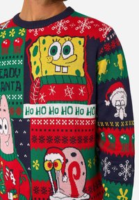 Pühadeteemaline kampsun, mis sisaldab värvikaid mustreid, sealhulgas tegelasi ja pidulikke motiive punases, rohelises ja tumesinis. Valmistatud kootud kangast.