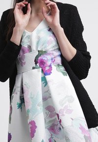 Robe florale blanche avec des aquarelles violettes et vertes, tissu texturé, taille ajustée, et un gilet tricoté noir. Mains positionnées près du visage.