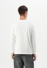 Young man wearing a plain white long-sleeve shirt and dark gray jeans facing away against a white background.