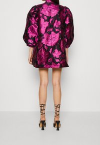 Kvinna som bär en svart miniklänning med stora glänsande rosa blommönster och puffärmar, kombinerat med svarta snörda högklackar.