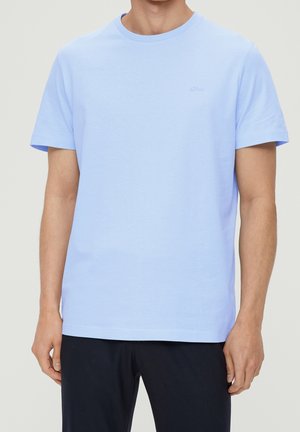 Man draagt een effen lichtblauw T-shirt met korte mouwen en ronde hals, en donkere broek; staat met ontspannen armen langs het lichaam.