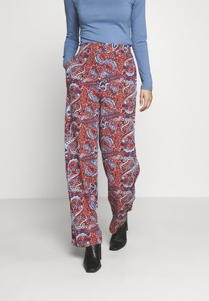 Femme portant un pantalon large rouge à motif paisley, un haut bleu à manches longues et des bottines noires, debout les mains dans les poches.