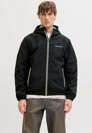 Junger Mann mit hellbraunen Haaren, trägt eine schwarze Kapuzenjacke mit Reißverschluss, darunter ein weißes Hemd und ausgewaschene graue Jeans, steht vor einem schlichten Hintergrund.