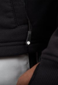 Čierna mikina na zips s textúrovanou tkaninou a malým embosovaným logom na zipsovom ťahadle. Interiér je v svetlejšej farbe.