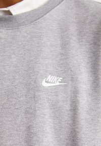 Siv sweatshirt iz bombaža z okroglim izrezom, rebrastim ovratnikom in belim vezena logotipom Nike na prsih. Mehka tekstura in sproščen dizajn.