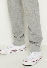 Jambes portant un pantalon de survêtement gris rentré dans des baskets en toile blanches avec des bandes rouges et noires sur les semelles, debout sur un sol clair.