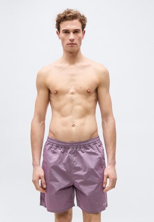 Giovane uomo con capelli ricci in piedi di fronte, indossa pantaloncini da bagno viola su uno sfondo grigio chiaro uniforme.