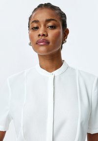 Witte blouse met een mandarin kraag, korte mouwen, een knoopsluiting en een textuurstof langs het voorpaneel. Glad, lichtgewicht materiaal.