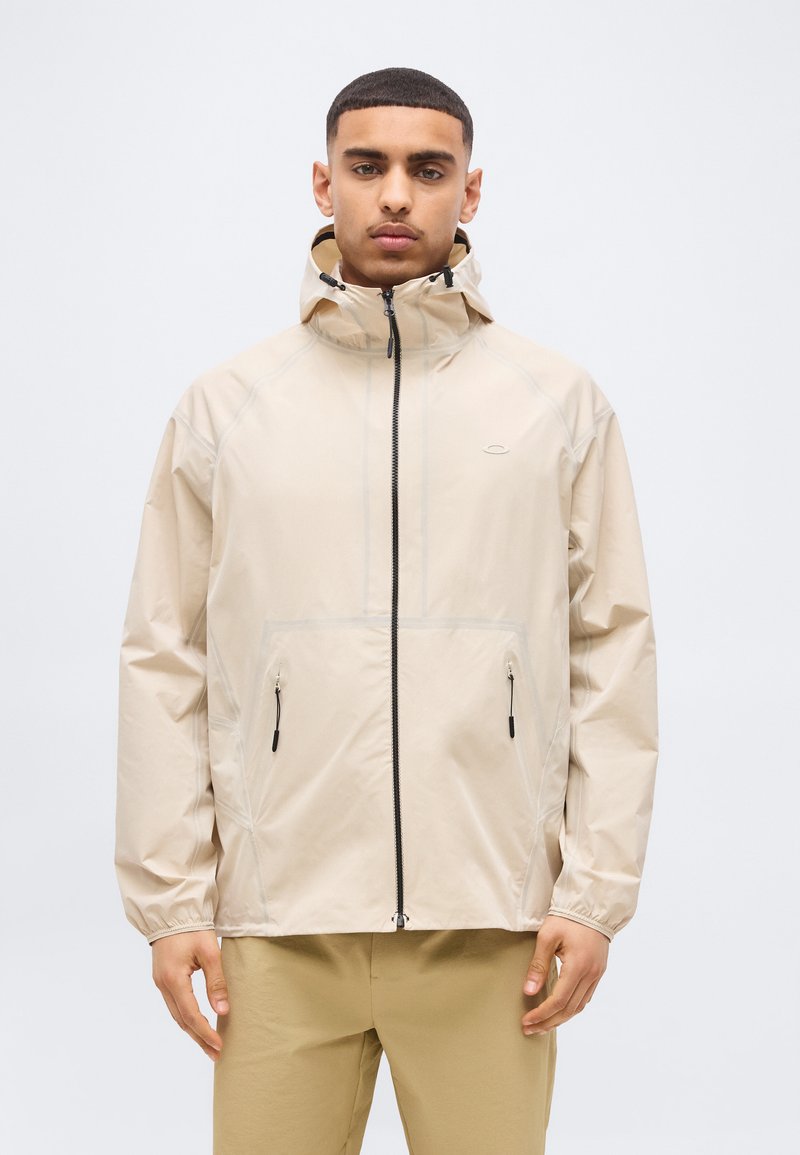 Άντρας που φοράει μπεζ μπουφάν με κουκούλα, με φερμουάρ στο μπροστινό μέρος και τσέπες, στέκεται μπροστά σε ένα απλό φωτεινό φόντο.