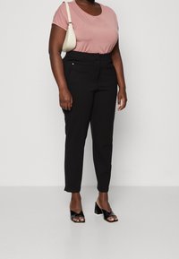 Pantalon noir à coupe droite avec poches avant, associé à un t-shirt rose pâle. Les chaussures sont des sandales à talons ouverts noires.