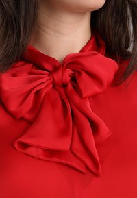 Blouse rouge avec un grand nœud doux à col, en tissu léger à texture lisse sans motifs visibles.