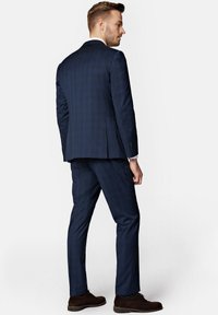 Granatowy garnitur w kratę o dopasowanym kroju. Posiada kurtkę z podwójnym rozcięciem, dopasowane spodnie oraz brązowe buty. Gładka tekstura tkaniny.