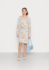 Vestido floral azul-claro com mangas longas, decote em V e cintura ajustada. Combinado com uma bolsa texturizada azul-claro e sandálias de tiras creme.