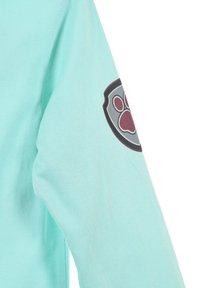 Mintgrünes Langarmshirt mit einem Patch, der ein graues Schild und einen burgunderfarbenen Pfotenabdruck auf dem linken Ärmel zeigt. Weicher Stoff mit glatter Textur.
