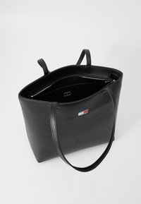 Schwarze Leder-Tasche mit zwei Griffen, einem Reißverschluss-Innenfach, glatter Textur und einem kleinen Logo-Patch in den Farben Rot, Weiß und Blau.