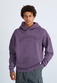 Lila Kapuzenpullover aus weichem Stoff, mit einem Frontlogo, lockerer Passform, rohen Kanten und einer geräumigen Kapuze, kombiniert mit grauen Hosen.