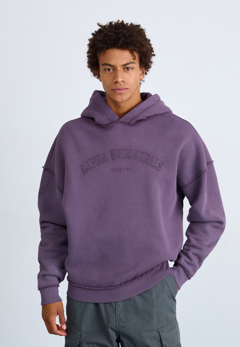 Jeune homme aux cheveux bouclés portant un sweat à capuche violet Alpha Industries et un pantalon cargo sombre, se tenant avec une expression neutre.