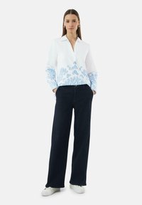 Witte shirt met blauwe geborduurde patronen op de mouwen, gecombineerd met donkerblauwe wijde broek en witte sneakers. Soepele stoftextuur.