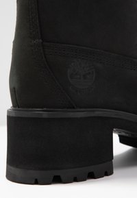 Botín de ante negro con un acabado texturizado, que presenta un tacón robusto y un logo grabado en el lateral, destacando los detalles de costura resistentes.